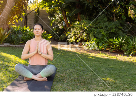 Asian woman practicing yoga cross?legged on yoga mat in home garden, showcasing dappled sunlight Asian woman practicing yoga cross?legged on yoga mat in home garden, showcasing dappled sunlight 128740150