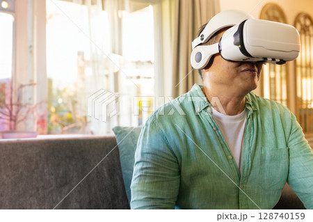 Asian senior man using VR headset on gray sofa near windows in living room, copy space Asian senior man using VR headset on gray sofa near windows in living room, copy space 128740159