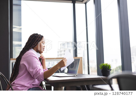 Woman reviewing documents and referencing laptop at table in office with headphones, copy space Woman reviewing documents and referencing laptop at table in office with headphones, copy space 128740177