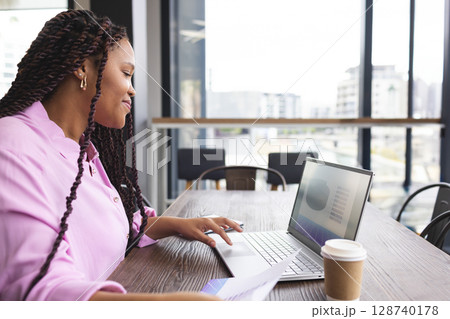 Woman typing on laptop at modern coworking space, holding document and coffee cup Woman typing on laptop at modern coworking space, holding document and coffee cup 128740178