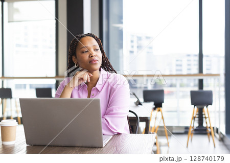 Woman sitting at table in modern coworking space, gazing thoughtfully with laptop and coffee cup 128740179
