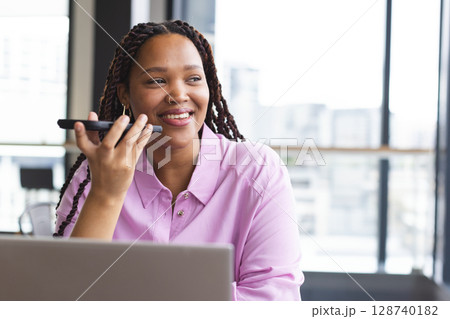 Diverse woman speaking into smartphone, working on laptop in office, with city skyline, copy space Diverse woman speaking into smartphone, working on laptop in office, with city skyline, copy space 128740182