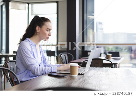 Woman typing on silver laptop at modern cafe with large windows, holding coffee cup and smartphone Woman typing on silver laptop at modern cafe with large windows, holding coffee cup and smartphone 128740187