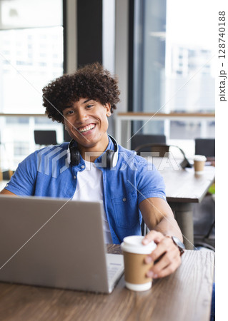 Young man working on laptop at modern cafe table, with headphones and holding paper coffee cup 128740188