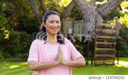 Woman in her 50s standing on lawn in backyard garden, holding hands in yoga greeting 128740202