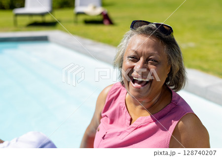 Relaxed senior woman enjoying sun on pool edge in backyard with sunglasses, copy space 128740207