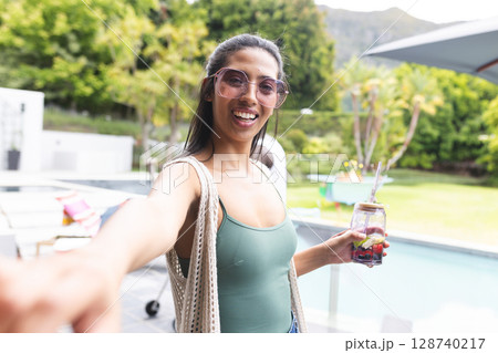 Asian woman standing at backyard pool smiling at camera, holding mason jar drink, carrying tote 128740217