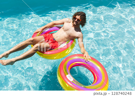 Young man floating on multicolored inflatable ring in outdoor swimming pool, holding second ring Young man floating on multicolored inflatable ring in outdoor swimming pool, holding second ring 128740248