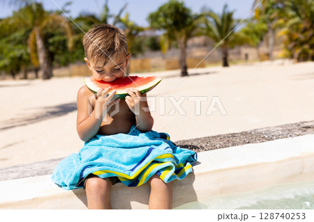 4-year-old boy sitting on pool edge at outdoor pool eating watermelon slice, with striped towel 128740253