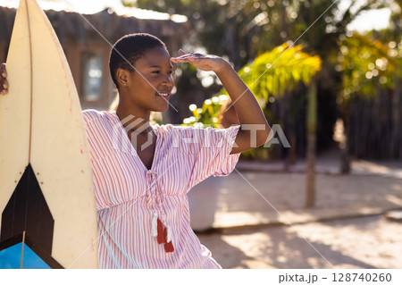 African American woman shading eyes while holding surfboard on sandy beach, gazing toward horizon 128740260