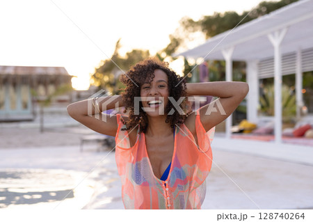 African American woman raising arms on boardwalk at sunset, wearing orange cover-up and bikini top 128740264