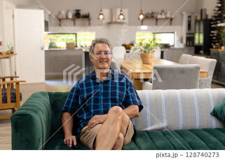 Senior man sitting cross-legged on green velvet couch in open-plan home, showing relaxed smile 128740273