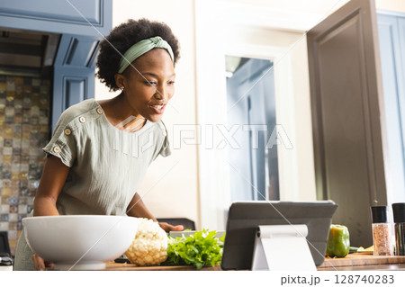Young African American woman reading recipe on tablet at wooden island, mixing vegetables in bowl Young African American woman reading recipe on tablet at wooden island, mixing vegetables in bowl 128740283