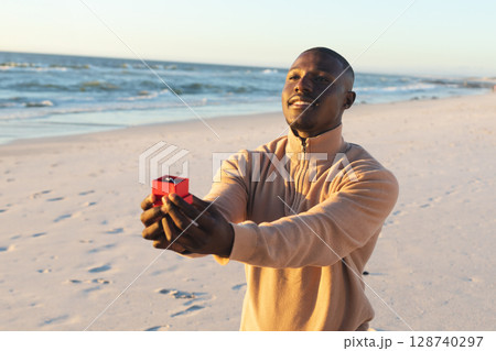 African American man kneeling on sandy shore, presenting red box holding diamond ring, copy space 128740297