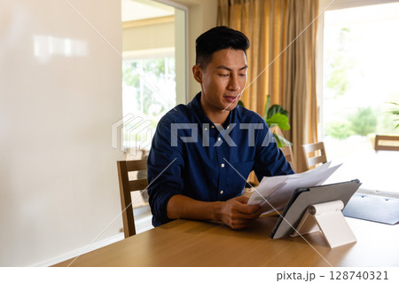 Young Asian man reviewing papers at wooden table in home office, with tablet and potted plant Young Asian man reviewing papers at wooden table in home office, with tablet and potted plant 128740321