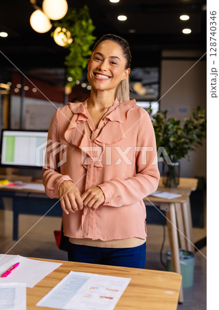 Woman in 30s standing behind wooden desk in modern office, smiling and reviewing charts Woman in 30s standing behind wooden desk in modern office, smiling and reviewing charts 128740346
