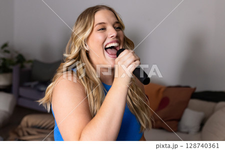 Young woman singing into black microphone in modern living room, with neutral-toned sofa 128740361