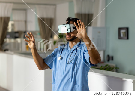 Male nurse using virtual reality headset and raising hands at reception desk, with stethoscope Male nurse using virtual reality headset and raising hands at reception desk, with stethoscope 128740375
