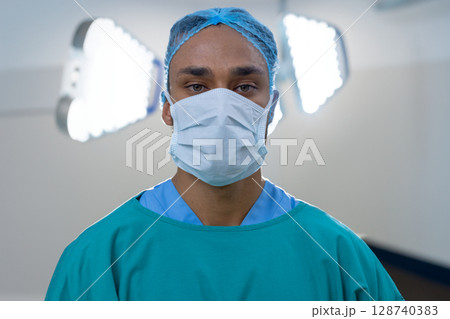 Male professional standing in operating room under LED lights, wearing surgical gown, cap and mask 128740383
