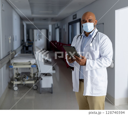 Male doctor reviewing tablet in hospital corridor, showing stethoscope and gurney, copy space Male doctor reviewing tablet in hospital corridor, showing stethoscope and gurney, copy space 128740394