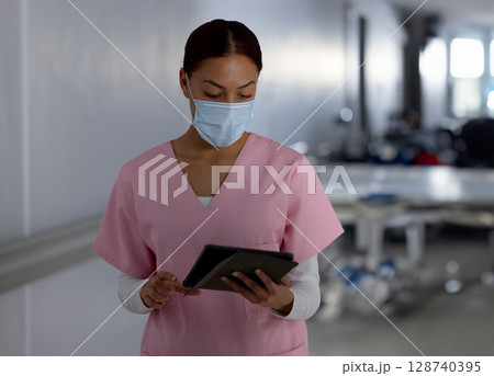 Female healthcare worker in pink scrubs studying tablet in hospital hallway wearing surgical mask Female healthcare worker in pink scrubs studying tablet in hospital hallway wearing surgical mask 128740395