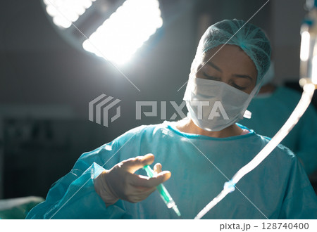 Young female surgeon with mask preparing syringe under surgical light with tubing in operating room 128740400