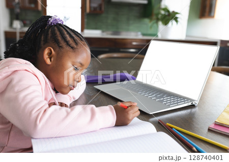African American child writing at kitchen table with red pencil, open notebook, laptop, copy space 128740401