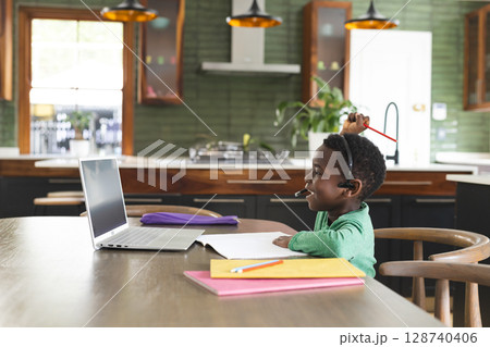 African American boy working on laptop in kitchen at table, with notebooks and headset, copy space African American boy working on laptop in kitchen at table, with notebooks and headset, copy space 128740406