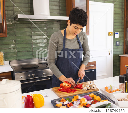 woman chopping red bell peppers on wooden cutting board in home kitchen, with spice jars woman chopping red bell peppers on wooden cutting board in home kitchen, with spice jars 128740423
