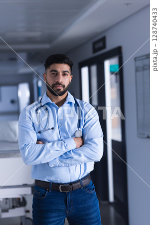 Indian male doctor standing with crossed arms in hospital hall, with stethoscope, gurney Indian male doctor standing with crossed arms in hospital hall, with stethoscope, gurney 128740433