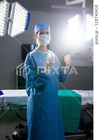 Female surgeon standing in operating room under overhead LED lights beside green draped table Female surgeon standing in operating room under overhead LED lights beside green draped table 128740438
