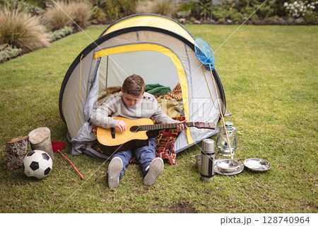 Child boy sitting at backyard tent, strumming guitar with soccer ball and butterfly net nearby 128740964