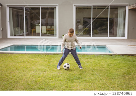 young boy dribbling soccer ball on backyard lawn in front of modern house, featuring pool 128740965