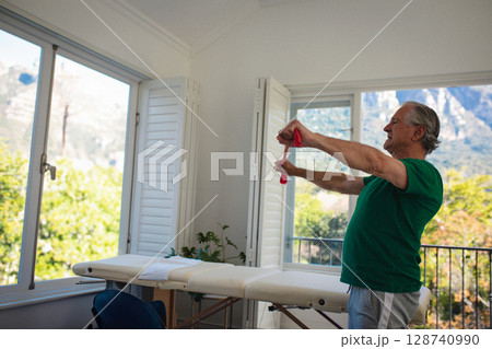 Senior man stretching red resistance band across chest in therapy room with shutters, copy space 128740990