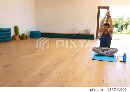 Man stretching arms overhead cross?legged on blue yoga mat in yoga studio with blocks, copy space Man stretching arms overhead cross?legged on blue yoga mat in yoga studio with blocks, copy space 128741001
