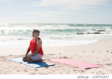 Senior man kneeling on blue yoga mat at sandy beach near shoreline rocks, copy space 128741017