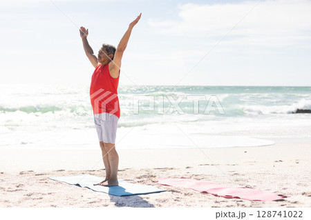 Senior man stretching overhead on light-blue yoga mat on beach, with red mat beside, copy space 128741022