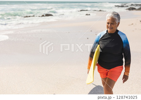 Senior woman walking along beach shoreline, carrying yellow surfboard, copy space Senior woman walking along beach shoreline, carrying yellow surfboard, copy space 128741025