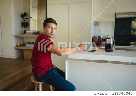 Man sitting on wooden stool at kitchen island, holding coffee mug with French press and smartphone 128741040