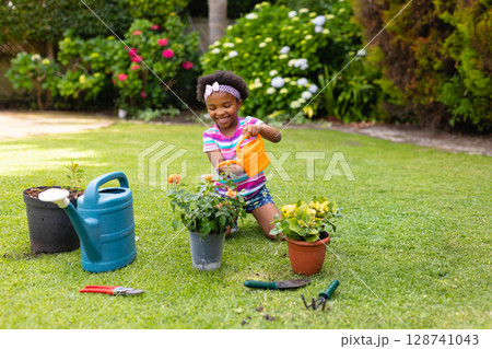 African American girl kneeling and watering plants in backyard garden, with watering cans and tools 128741043
