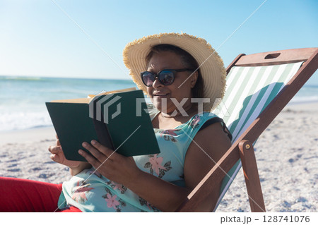 Senior woman reading book in deck chair on beach wearing sun hat and sunglasses, copy space Senior woman reading book in deck chair on beach wearing sun hat and sunglasses, copy space 128741076