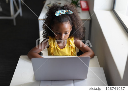 Child using silver laptop at white desk in study, with open notebook and decorative hair bow 128741087
