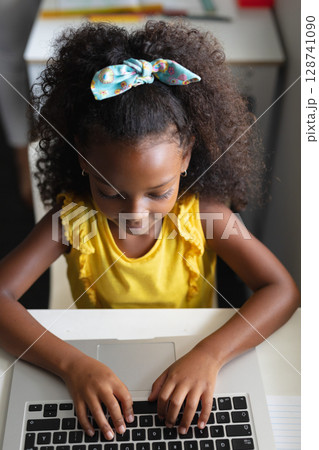 Child girl typing on silver laptop at home desk, showing focus with notebook and hair bow 128741090