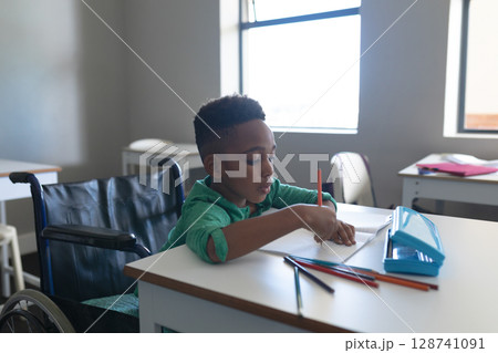 African American boy writing in notebook at wheelchair desk in classroom, using colored pencils 128741091