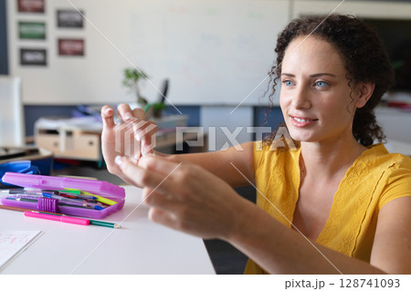 Female instructor examining small object at desk in classroom, with pink pencil case, copy space Female instructor examining small object at desk in classroom, with pink pencil case, copy space 128741093
