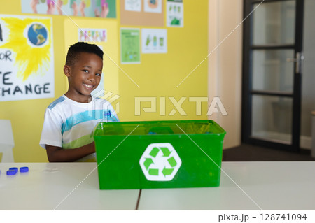 African American boy placing clear plastic bottle into green recycling bin in classroom, copy space African American boy placing clear plastic bottle into green recycling bin in classroom, copy space 128741094
