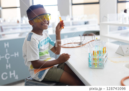 African American boy wearing goggles holding orange test tube in lab with tube rack, copy space African American boy wearing goggles holding orange test tube in lab with tube rack, copy space 128741096