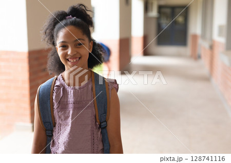 Female child student standing in school walkway with brick columns, windows, wearing blue backpack 128741116