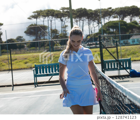 Woman walking alongside tennis net on outdoor tennis court, holding tennis racket 128741123