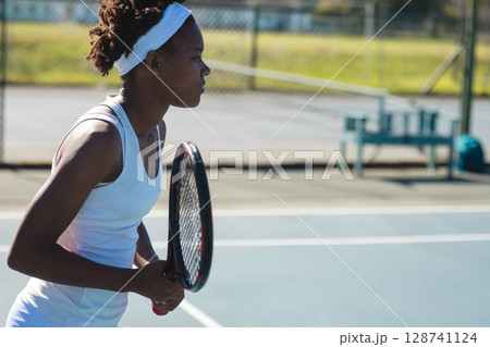 bi-racial young adult woman holding tennis racket ready on tennis court with headband, copy space 128741124